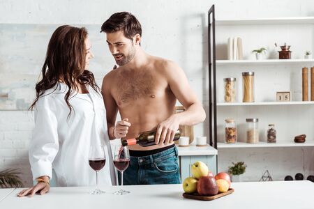 Handsome and shirtless man pouring red wine in wine glasses near attractive girl in kitchenの写真素材