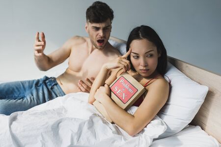 Selective focus of offended Asian woman holding problems in sex book while lying near quarreling boyfriendの写真素材