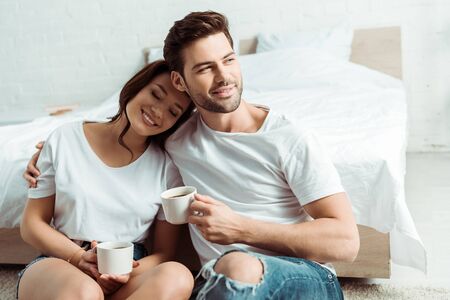 Happy man sitting with cheerful girl and holding cup in bedroomの写真素材