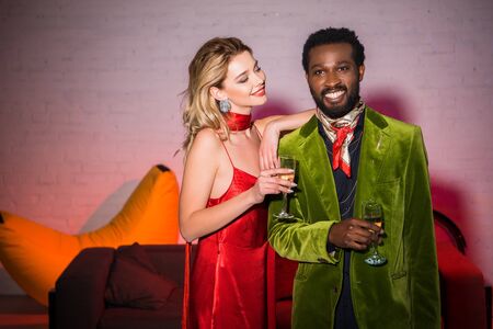 Cheerful and attractive woman in red dress looking at handsome African American man with champagne glassの写真素材