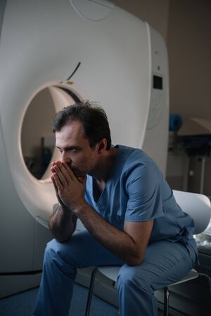 Thoughtful radiologist sitting near computed tomography scanner in hospital and looking awayの写真素材