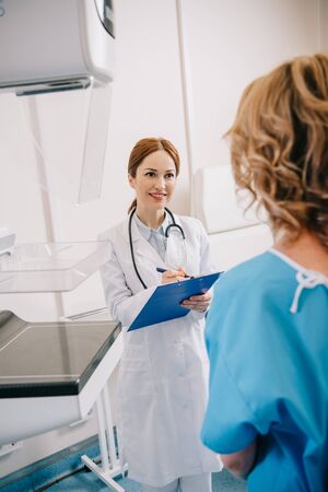 Selective focus of smiling radiographer writing on clipboard near patient in clinicの写真素材