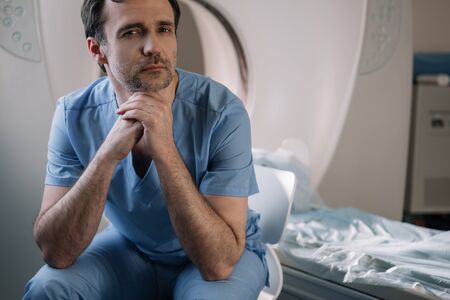 Pensive doctor sitting near computed tomography scanner in hospital and looking at camera