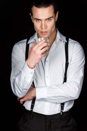 Handsome man in white shirt and suspenders holding glass of whiskey while looking at camera isolated...