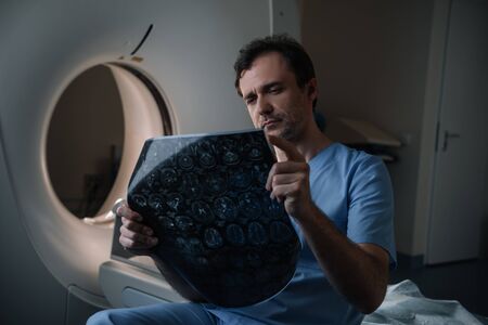 Attentive doctor looking at tomography diagnosis while sitting near ct scannerの写真素材