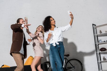 Low angle view of attractive African american girl taking selfie with friends holding champagne glassesの写真素材