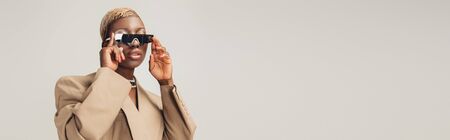 Fashionable African American woman posing in sunglasses and beige jacket isolated on grey backgroundの写真素材