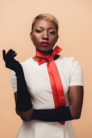Young African American woman posing in white dress, red scarf and black gloves isolated on beige backgroundの写真素材