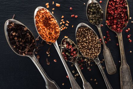 top view of pepper, candied fruit, coriander and carnation seeds in silver spoons on black textured...