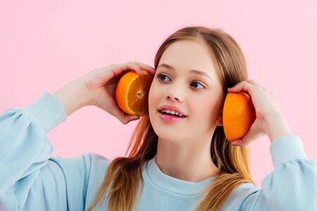 smiling pretty teenage girl holding orange halves near ears isolated on pink
