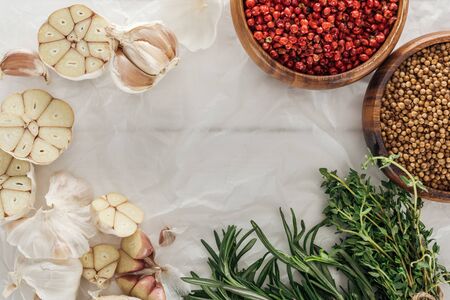 top view of garlic cloves, pink peppercorn and coriander in bowls, rosemary and thyme on white paper...