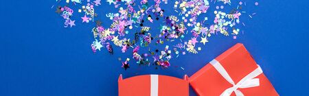Panoramic shot of red gift box and multicolored confetti on blue background, surprise conceptの写真素材