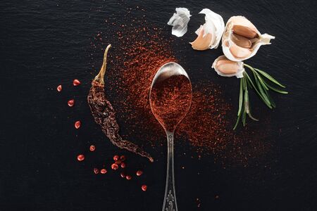 top view of red pepper powder in silver spoon on black background with dried chili peppe, herbs and...