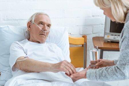 cropped view of senior woman giving glass of water to sick husband in hospitalの写真素材