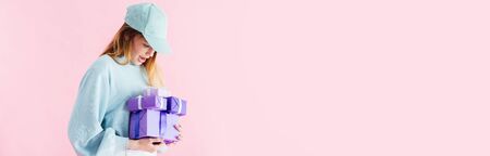 panoramic shot of excited pretty teenage girl in cap looking at purple gifts isolated on pink