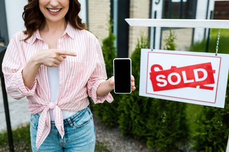 Cropped view of cheerful woman pointing with finger at smartphone with blank screen