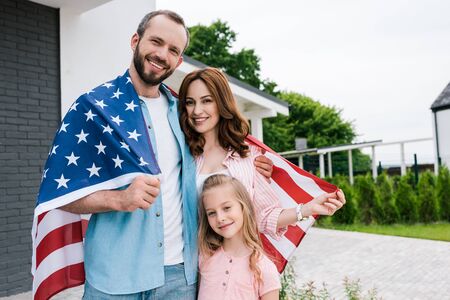 Happy kid near cheerful parents with American flag standing near houseの写真素材