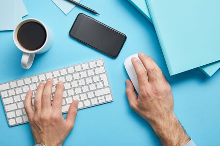Cropped view of man using computer keyboard and computer mouse at workplace with papers, smartphone...