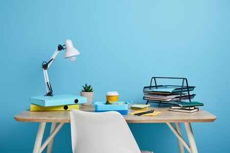 Workplace with papers and lunch box with coffee in plastic cup on wooden table on blue backgroundの写真素材