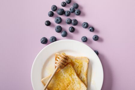 Top view of toasts with honey, wooden dipper on white plate and scattered blueberries on violet backgroundの写真素材
