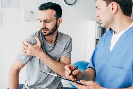 Selective focus of patient touching his shoulder and suffering from pain and doctor writing...