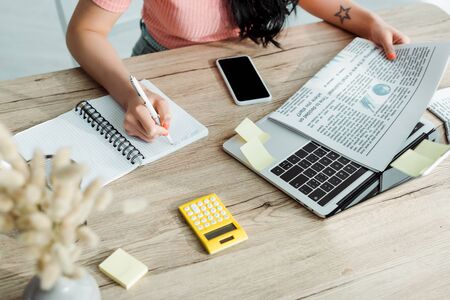 Cropped view of young woman writing in notebook near newspaper and gadgetsの写真素材