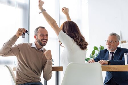 Selective focus of cheerful man holding car keys and gesturing near woman and car dealer