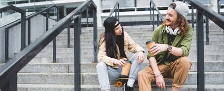 Low angle view of happy couple sitting on skateboard with paper cup in hands, looking at each otherの写真素材