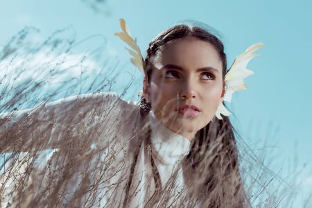 Low angle view of elegant woman in white swan costume standing near dry bush, looking away