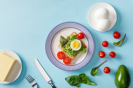 Top view of toast with guacamole, avocado, boiled eggs, butter, scattered cherry tomatoes and...