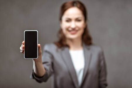Selective focus of cheerful businesswoman holding smartphone with blank screen on grey background