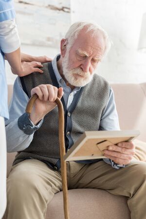 Nurse standing near upset grey haired man with photo frame, putting hands on his shoulder
