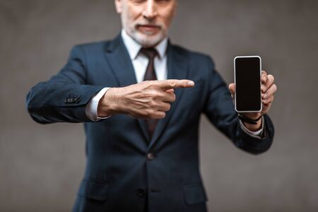 Selective focus of businessman pointing with finger at smartphone with blank screen on grey backgroundの写真素材