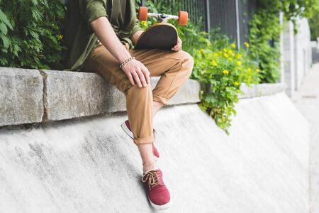 Cropped view of man in casual wear holding skateboard in hand, sitting on borderの写真素材