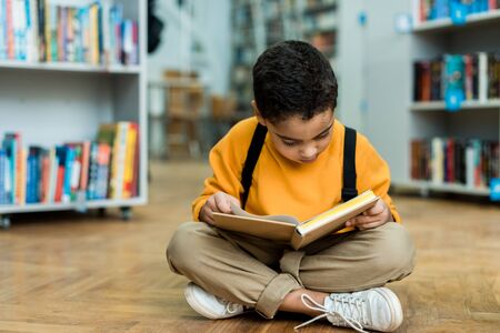 Cute African American boy sitting on floor and reading bookの写真素材