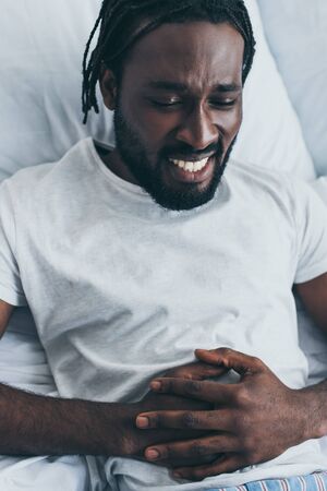 Exhausted African American man suffering from stomach pain in bedroomの写真素材