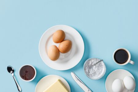 Top view of fresh boiled eggs, butter, jam on white plates, yogurt with chia seeds, coffee, spoon and knife on blue backgroundの写真素材