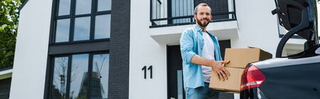 Panoramic shot of happy man holding boxes near car and modern houseの写真素材