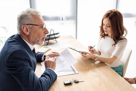 Car dealer in glasses looking at attractive woman with clipboard and penの写真素材