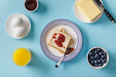Top view of toasts with jam, orange juice, boiled eggs, butter and blueberries on blue backgroundの写真素材