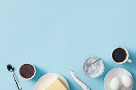 Top view of butter, jam, yogurt with chia seeds, coffee and fresh eggs on white plates, knife and...