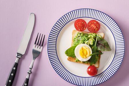 Top view of toast with guacamole, boiled egg, spinach, cherry tomatoes, fork and knife on ornamental...
