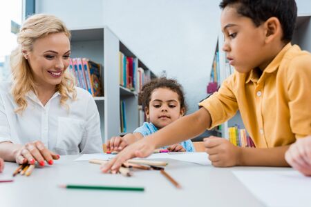 Selective focus of attractive and happy woman looking at cute African American kid taking color pencilの写真素材