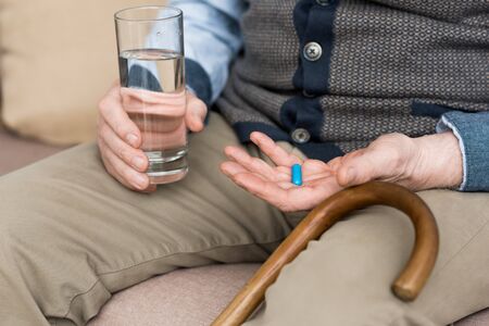 Cropped view of elderly man holding pill and glass with water in hands, sitting on couchの写真素材