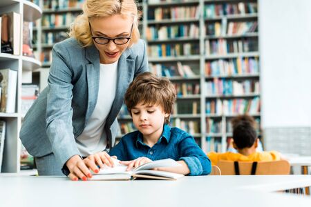 Selective focus of attractive woman in glasses standing near kid reading bookの写真素材