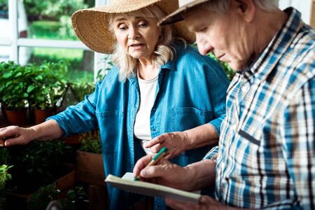 Selective focus of senior woman talking with husband writing in notebookの写真素材