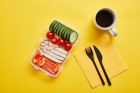 Top view of lunch box with chicken and fresh vegetables and cup of coffee on yellow background