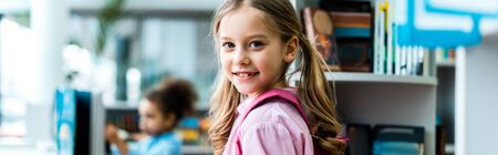 Panoramic shot of cheerful kid with pink backpack standing in library