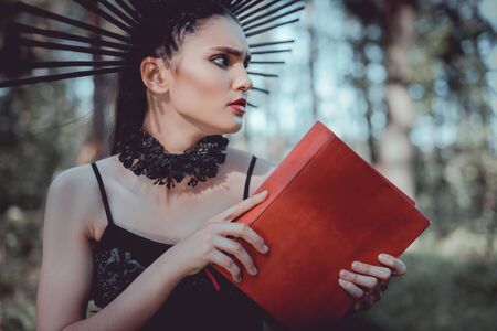 Beautiful woman in witch costume standing on forest background, holding red book, looking awayの写真素材
