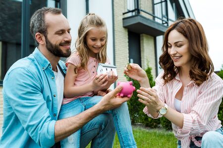 Happy woman putting golden coin in piggy bank near husband and daughterの写真素材